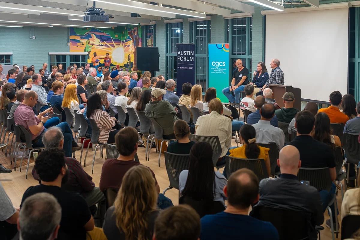 A large audience fills the room at an Austin Forum event, with a colorful mural on the left wall and panelists seated on stage beneath Austin Forum banners on the right